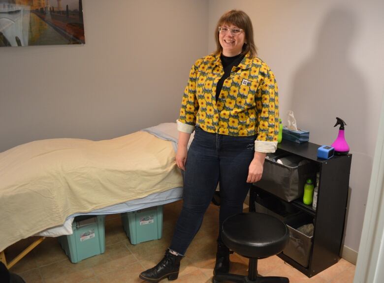 A woman with shoulder length brown hair stands in a room with a bookshelf and a massage bed. 