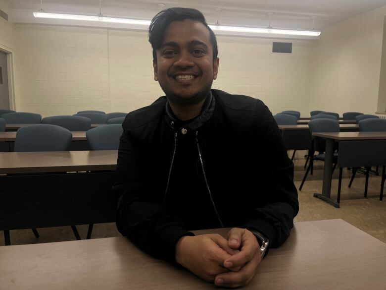 A man with short brown hair and a black jacket sits in a classroom with chairs and tables behind him. 