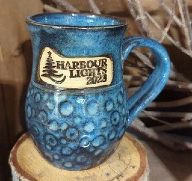 Blue clay mug that reads Harbour Lights 2023 in beige clay with a small Christmas tree design.