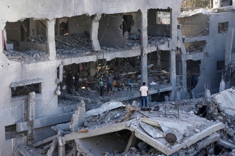 A man standing on rubble while two others speak to him from the opening of an opposite destroyed residential building.