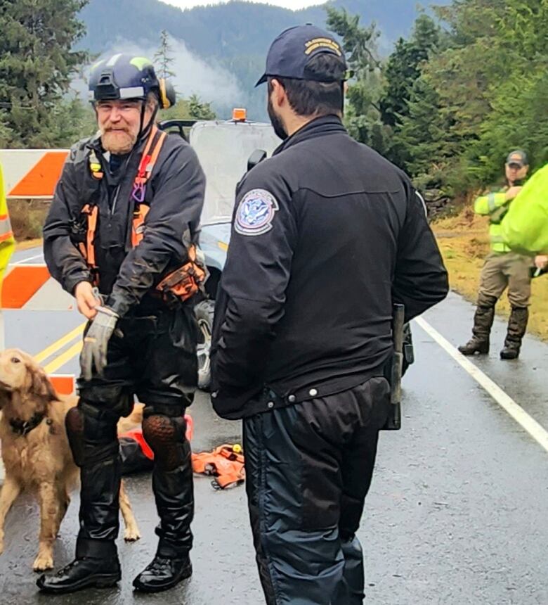 Two men in search and rescue gear stand with a dog on a road, with forest and mountains in the background.