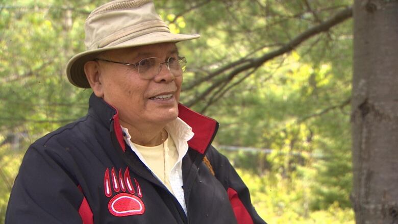 A man stands outdoor. He is wearing a yellow hat and a blue jacket. 