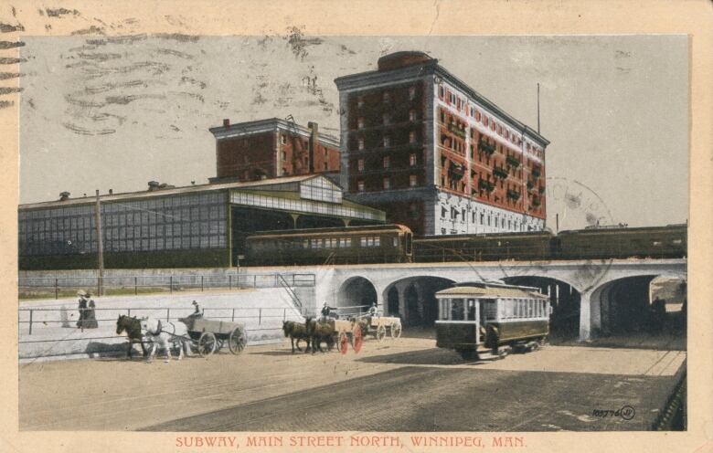 Black and white photo showing a streetcar and horse carts going under a bridge with trains above.