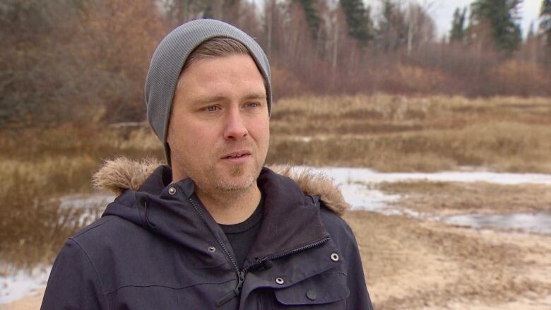 A man stands outside with grasslands and a forest behind him.