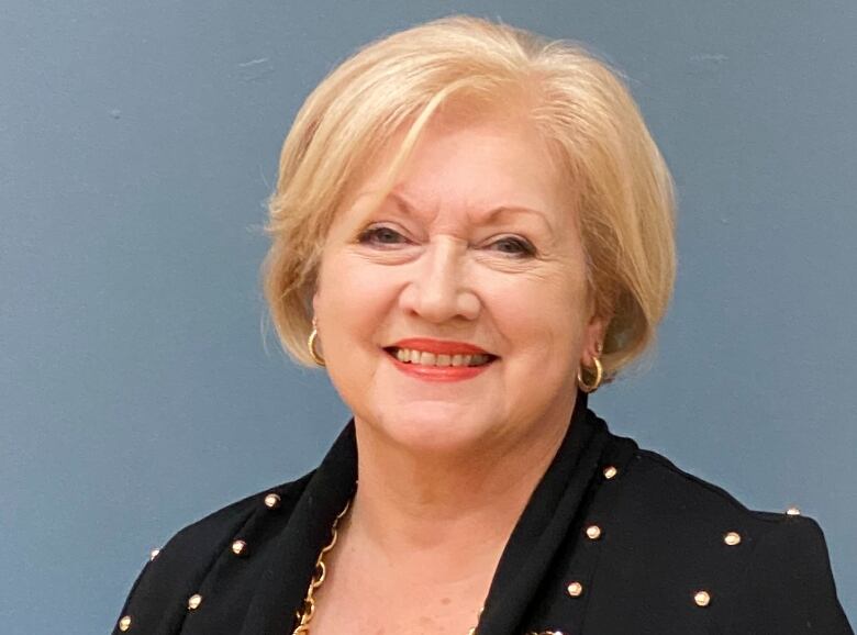 A woman with blonde hair and a navy blue blouse with gold buttons smiles.