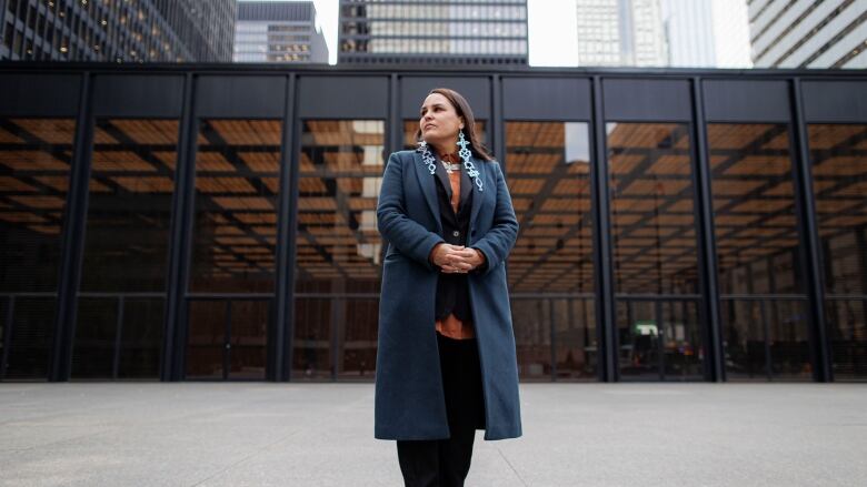 A woman with dark hair and dark jacket stands in front office building in downtown Toronto. Her orange blouse is offset by bold silver jewellry with Indigenous images.  