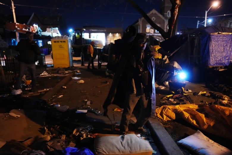 A person can be seen clearing an outdoor encampment, with debris on the ground.