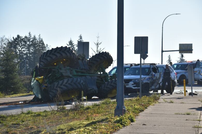 A green tractor lays on its side on a road, next to a mangled police car.