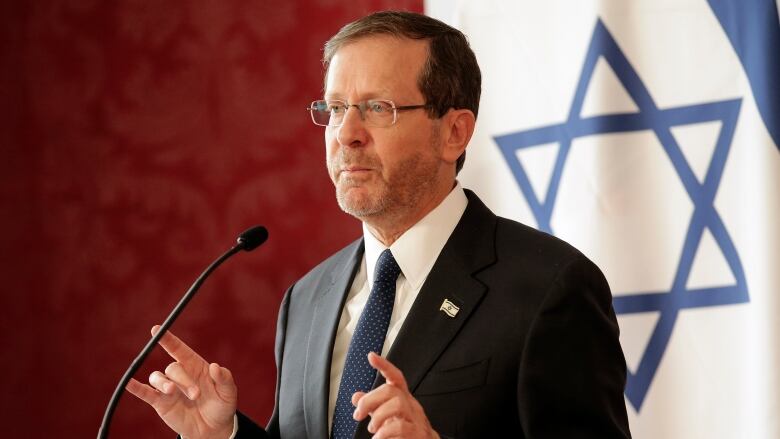 Issac Herzog, a white male wearing a suit, speaks from a podium. 