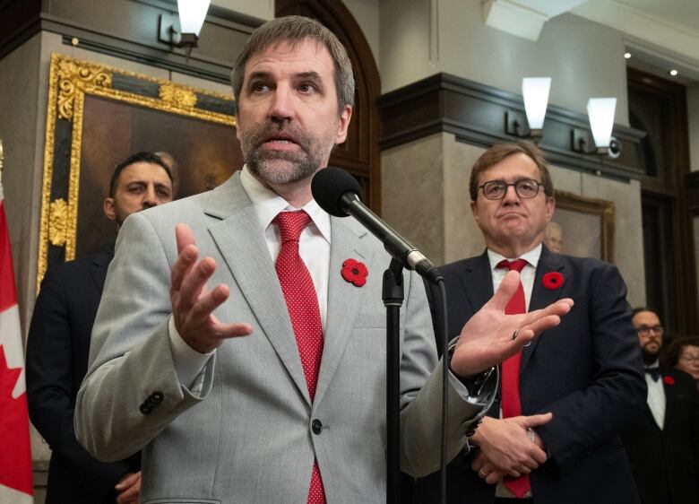 Men in suits at a news conference. A bearded man is taking with his hands raised.