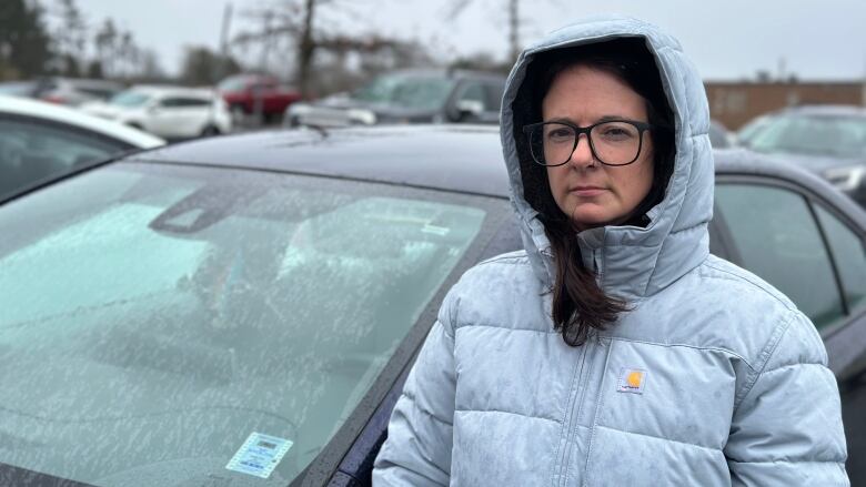 A woman wearing glasses stands by her car in a winter coat.