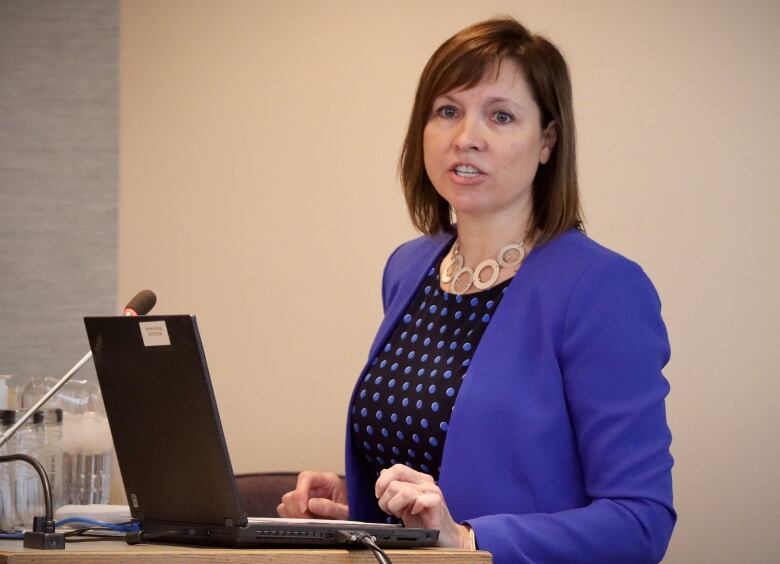 A woman in a blue jacket with a black blouse and blue polka dots stands and speaks in front of a computer and microphone.