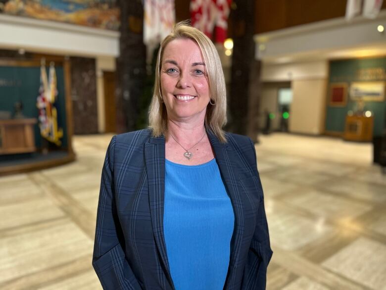 A smiling woman with blonde hair wearing a blue plaid blazer stands inside Confederation Building.