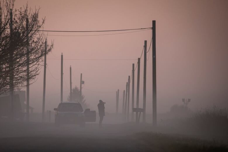 A woman shrouded in thick fog takes a picture next to a car.