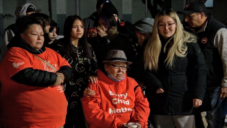 Two women in orange shirts, one standing, the other sitting, are pictured standing with supporters. One supporters has her hands on the shoulder of the woman who is sitting to comfort her. 