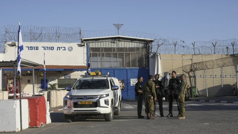 A group of armed, uniformed people stand by an official vehicle in front of a gate and walls with barbed wire.