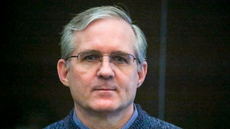 A bespectacled, clean-shaven man wearing a sweater and a collared shirt underneath is shown in a closeup photograph.