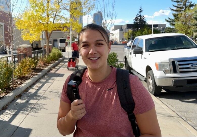 A woman wearing a T-shirt and a backpack stands on a city sidewalk on a sunny day.