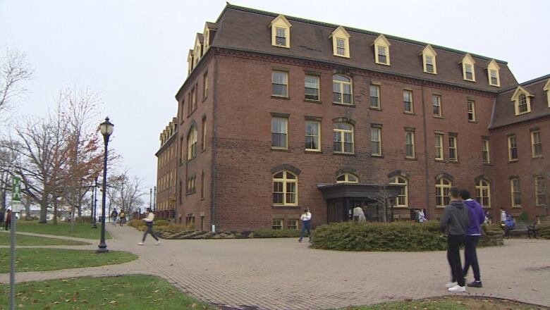 An old brick building with yellow window framing sits on a university quad, with students walking around. 