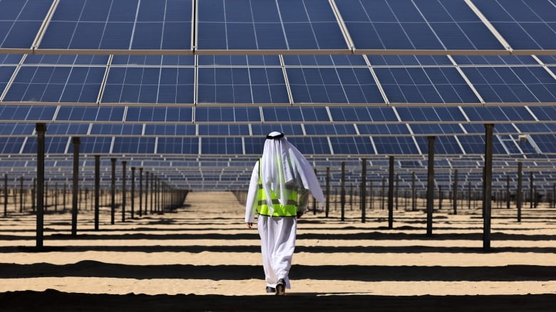 Man wearing a traditional Emirati white robe and head scarf with a yellow safety vest walks away from the camera, toward rows of solar panels