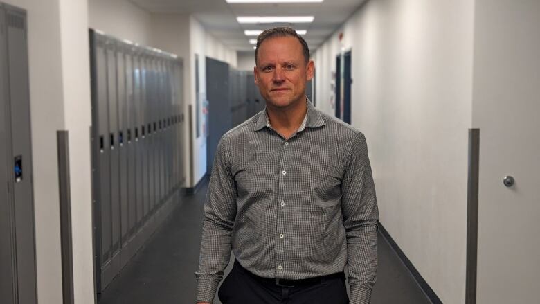 A man with a grey shirt stands in the middle of along hallway, with school lockers along the sides of the wall.