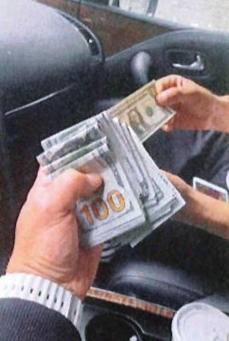 A stack of $100 US bills in someone's hands, on the driver side of a vehicle. 