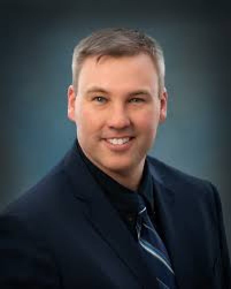 Smiling man with short brown-grey hair wears navy suit jacket, shirt and tie.