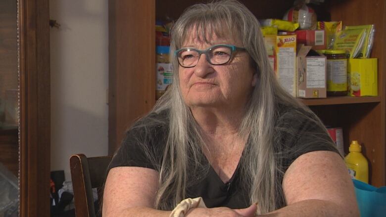 A woman with long grey hair and glasses sits at her kitchen table, with  canned goods on the shelves behind her. 