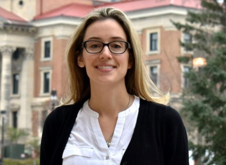 A woman with long hair and glasses stands on campus of the University of Manitoba.