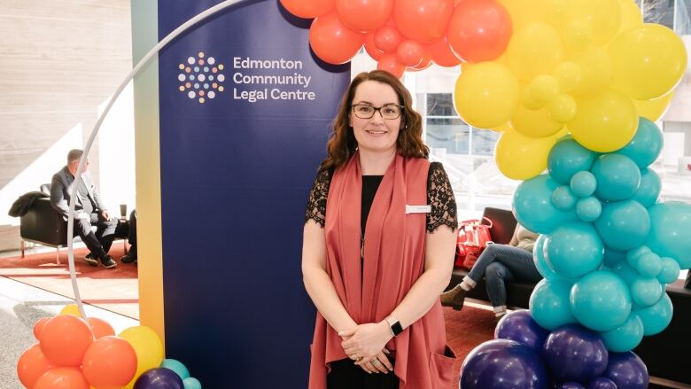 A woman with dark hair and glasses in front of a banner, surrounded by balloons. 