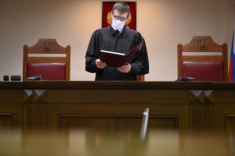 A Russian judge wearing a black robe and a face mask stands while reading a decision in a courtroom.