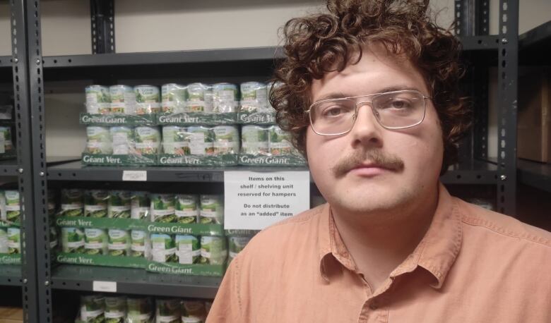 man with orange shirt and glasses standing in front of a shelf full of cans of beans