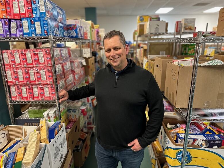 A man stand in an isle between shelves of donated, non-perishable food at a food bank.  He has short dark hair and wears a black sweater. 