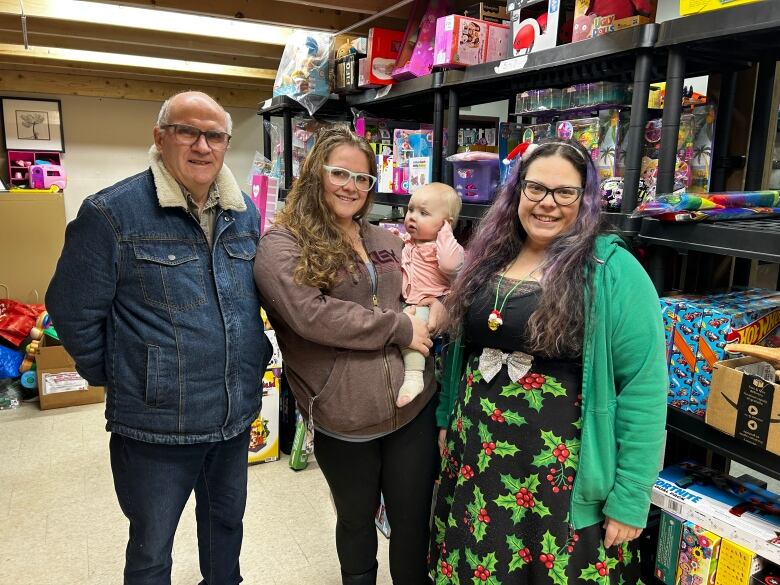 A man in a denim jacket stands next to a woman with long, curly brown hair holding a baby dressed in pink. She stands next to a smiling woman wearing a long skirt patterned with mistletoe.