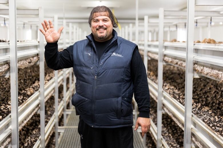 A farmer waves in a greenhouse-style building with racks of mushrooms.