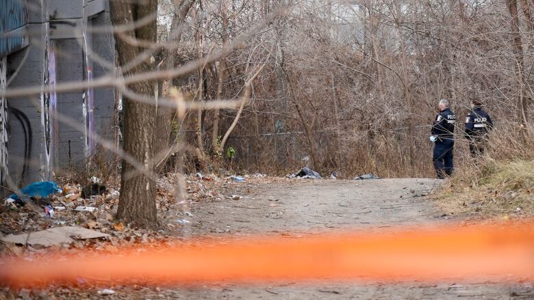 Orange police tape in foreground. Two officers in background look at debris on ground near dirt road.