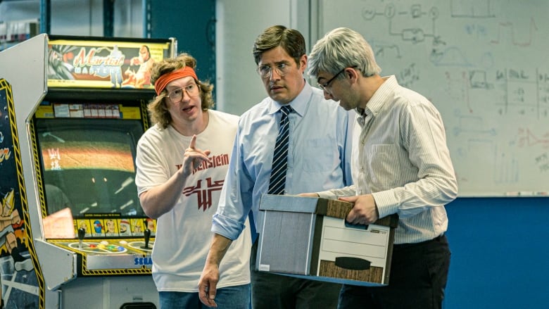 Matt Johnson talking to Rich Sommer and Jay Baruchel in a photo from TV series BlackBerry.  He's wearing a Wolfenstein T-Shirt and standing in front of an arcade machine.