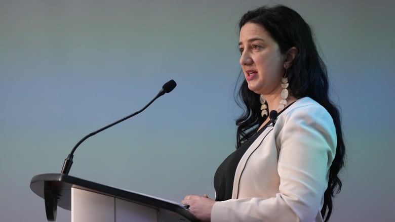 B.C. Human Rights Commissioner Kasari Govender is shown from the side as she speaks into a microphone at a lectern. Govender has long, dark, wavy hair and wears pale dangly earrings and a light-coloured blazer.