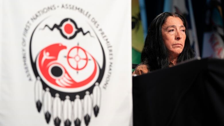 A woman sits on stage the background while the Assembly of First Nations logo is see out of focus in the foreground.