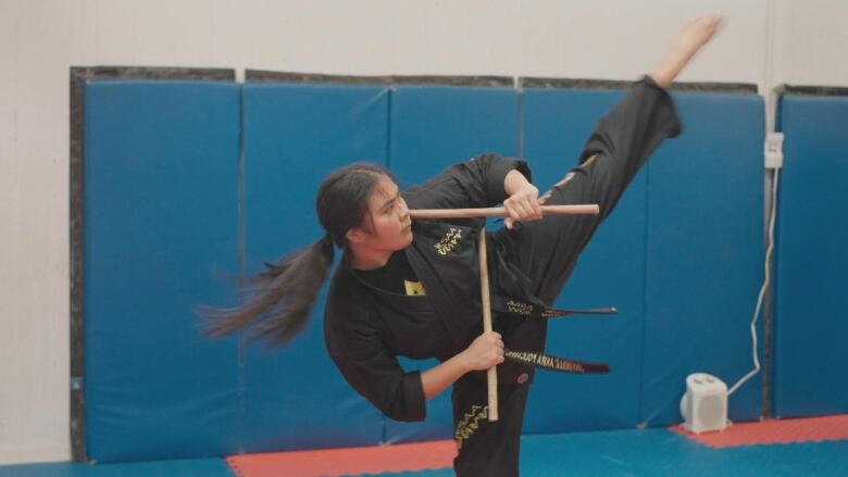 Young woman in a martial arts studio performs a sweeping high kick. She has long dark hair tied in a pony tail and is wearing a black karate uniform. 