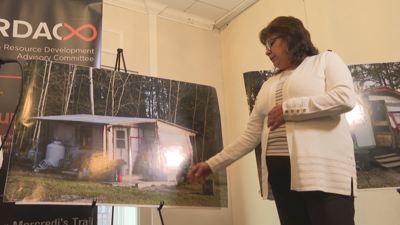 A middle-aged woman in a white cardigan and black pants pointing to a blown up image of her beat up house