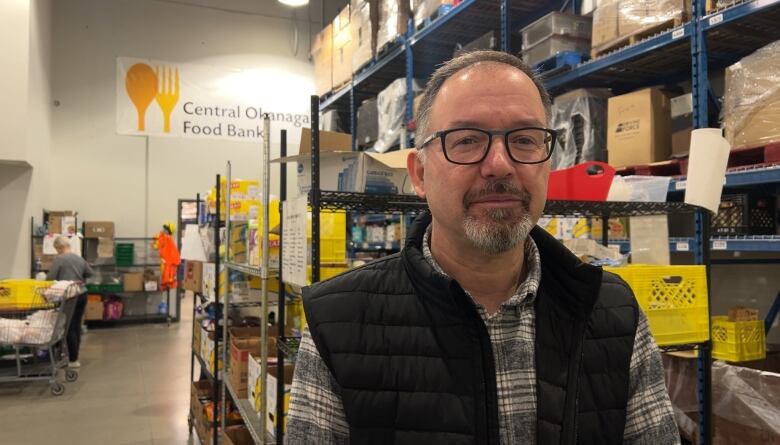 Trevor Moss is a middle aged man with black-framed glasses and a goatee and he is standing in the distribution center at the Central Okanagan Food Bank for an interview with the reporter. 