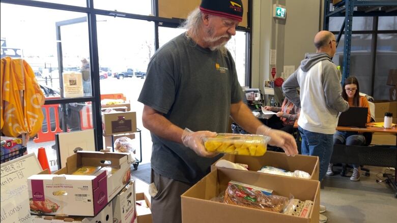 A middle-aged volunteer with a dark toque and a grey beard is loading a package of cut fruit into a cardboard food hamper.