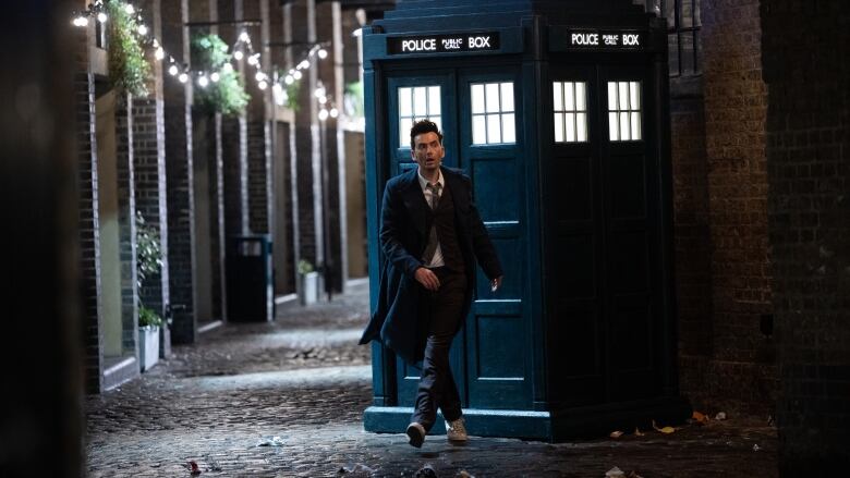 A man in a suit leaves a blue phone booth in an alley.