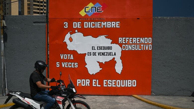 A helmeted motorcyclist rides by a wall bearing a poster that shows a map and a date of December 3rd for an upcoming vote.