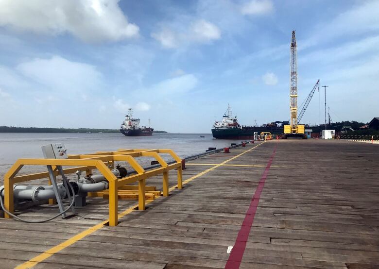 A wooden pier is shown in the foreground, and two ships and a crane in the background.