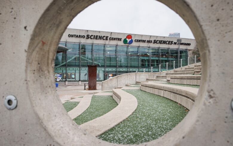 Exterior signage on a building saying 'Ontario Science Centre' in English and French is viewed through a round hole in a concrete wall.   