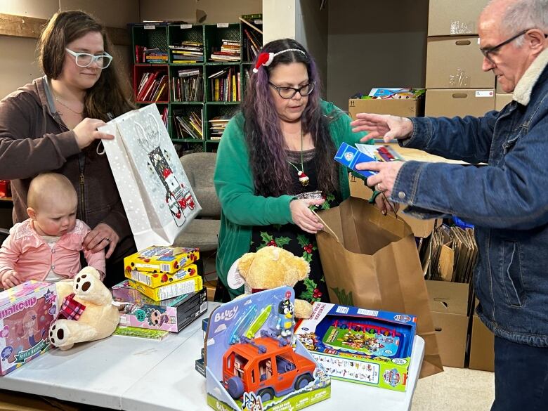 Two women and a man stand together over a table filled with toys, picking them up and placing them in bags. There's also a baby dressed in pink on the table.