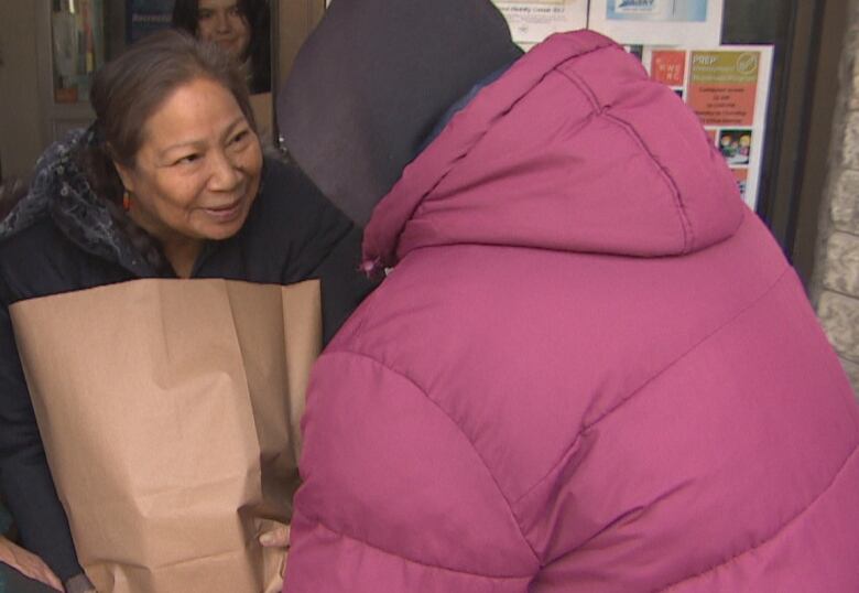 Food bank workers hand a paper bag of groceries to a female client wearing a pink jacket.