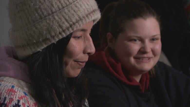 A woman in a toque and sweater has her profile facing the camera while her non-binary partner is smiling in the background.